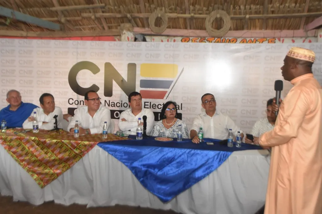 Magistrados del Consejo Nacional Electoral en la mesa de la sesión solemne en San Basilio de Palenque, mientras un hombre con atuendo tradicional y kufi les habla con un micrófono