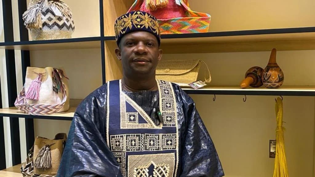 Pedro Adán Torres, con traje azul bordado y gorro tradicional, frente a una estantería de mochilas y artesanías colombianas