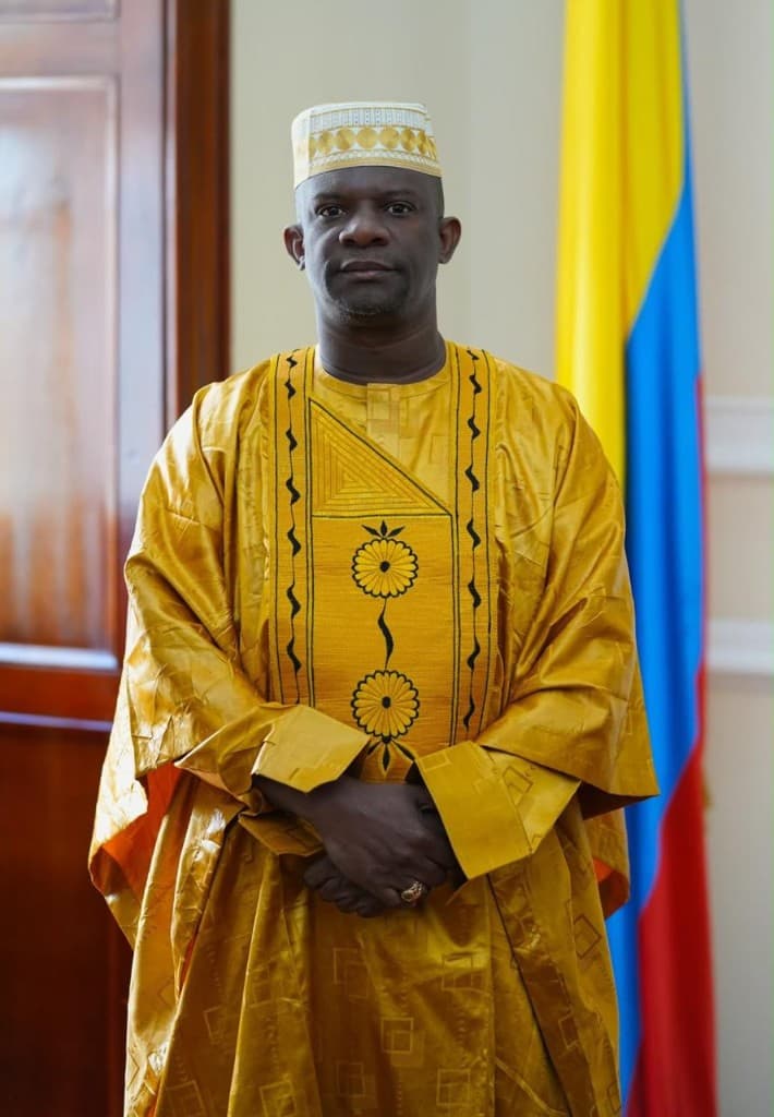 Pedro Adán Torres Pérez de pie, con traje tradicional dorado y kufi bordado, junto a la bandera de Colombia